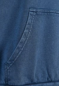 Tessuto in denim blu con una grande tasca frontale. La texture è liscia con una leggera lucentezza, che mette in evidenza le cuciture lungo i bordi.
