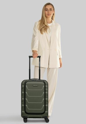 Femme en costume crème tenant la poignée d'une valise rigide verte à roulettes sur fond gris.