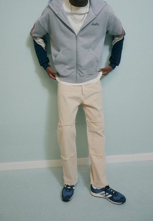 Sudadera gris con cremallera con acentos en azul y rojo, camiseta de color crema, pantalones desgastados en beige y zapatillas azul con acentos en negro. Fondo liso.