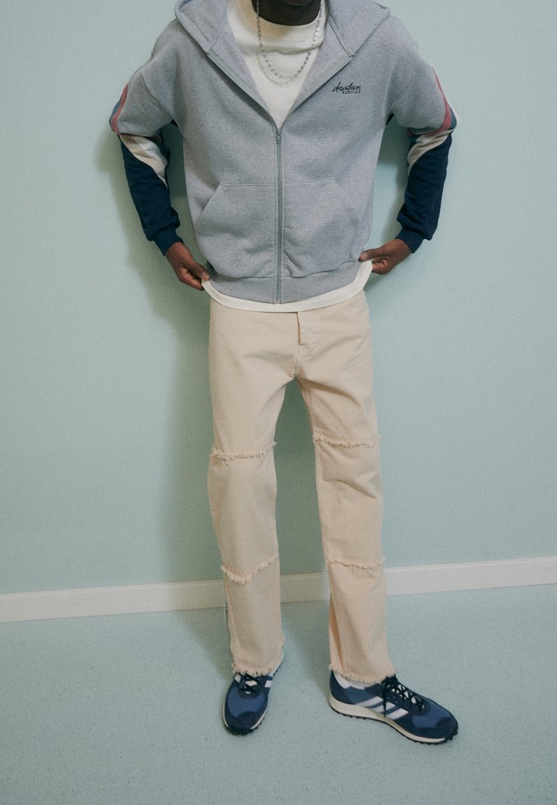 Felpa grigia con zip e dettagli blu e rossi, t-shirt crema, pantaloni beige strappati e sneakers blu con dettagli neri. Sfondo semplice.