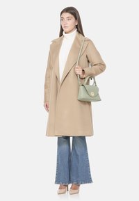 Manteau en laine beige avec de larges revers, associé à un col roulé blanc, un jean évasé en denim et un sac à main vert avec une fermeture dorée ronde.