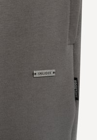 Tela gris con textura acanalada y una etiqueta metálica que dice "SMILODOX." Cuenta con una etiqueta negra con el nombre de la marca en el lateral.