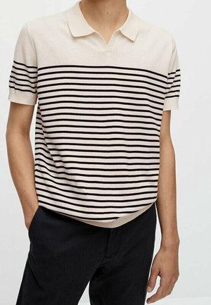 Homme portant un polo beige à manches courtes avec des rayures horizontales noires et un pantalon noir, debout avec une main dans la poche.