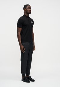 Άνδρας που στέκεται, φορώντας μαύρο πόλο με λευκό λογότυπο, σκουρόχρωμο καρό παντελόνι jogger και μαύρα δερμάτινα αθλητικά παπούτσια, μπροστά σε απλό ανοιχτό φόντο.