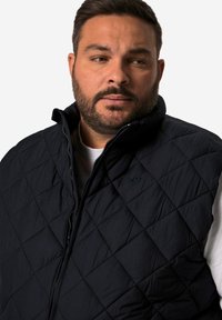 Prošívaná námořnická vesta s vysokým límcem, předním zipem a malým vyšitým logem. Materiál se zdá být lehký s texturovaným povrchem.