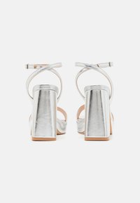Silver metallic high-heeled sandals feature a chunky rectangular heel, crossed ankle straps, and a smooth texture. Open back design.