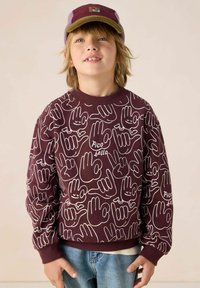 Sweat-shirt bordeaux avec motif graphique de mains blanches, présentant des poignets et un col côtelés. Coupe décontractée associée à un jean bleu clair.