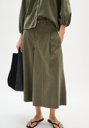 Oliwkowe spodnie culotte z zakładkami, wykonane z miękkiego materiału. W połączeniu z dopasowaną koszulą i czarną torbą typu tote. Na nogach japonki.
