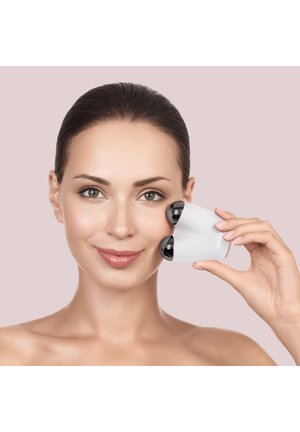 Femme utilisant un masseur facial manuel blanc avec deux rouleaux noirs sur sa joue droite sur un fond uni.