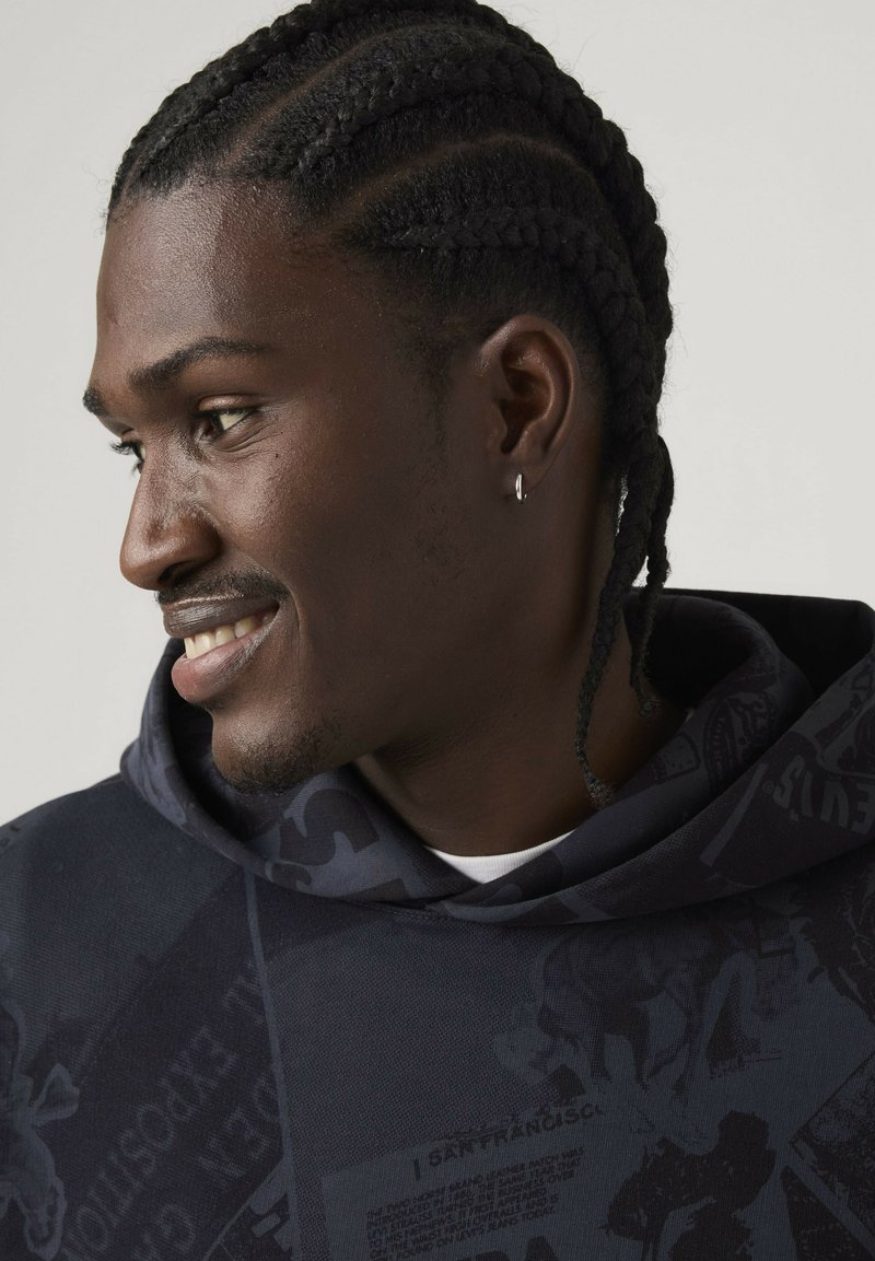 Man with braided hair and a small hoop earring smiles, wearing a dark hoodie with graphic text and patterns.
