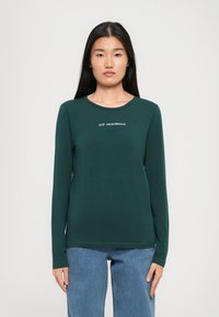 Kobieta w ciemnozielonej bluzie z długim rękawem z logo "EA7 Emporio Armani" i niebieskich dżinsach, stojąca na tle jasnego, jednolitego tła.