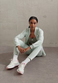 Light green athletic tracksuit with white stripes, fitted tank top, and white/pink sneakers. The fabric appears smooth and breathable.