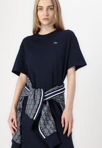 Navy cotton t-shirt with short sleeves, featuring a small green logo. A patterned navy and white sweater is tied around the waist.