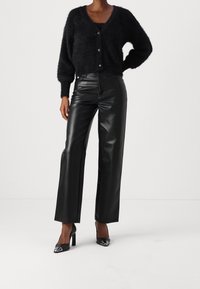 Czarny, puszysty kardigan z okrągłymi guzikami założony na czarną bluzkę; w połączeniu z wysokim stanem, błyszczącymi czarnymi spodniami z sztucznej skóry i czarnymi szpilkami.