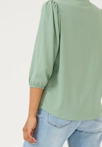 Blouse à manches bouffantes en tissu texturé vert clair avec un col rond et des poignets élastiques, portée avec un jean en denim bleu.