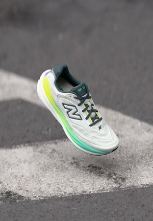 Zapatilla deportiva blanca y azul verdoso con cordones grises y suela con degradado neón suspendida sobre una carretera de asfalto con marcas blancas.