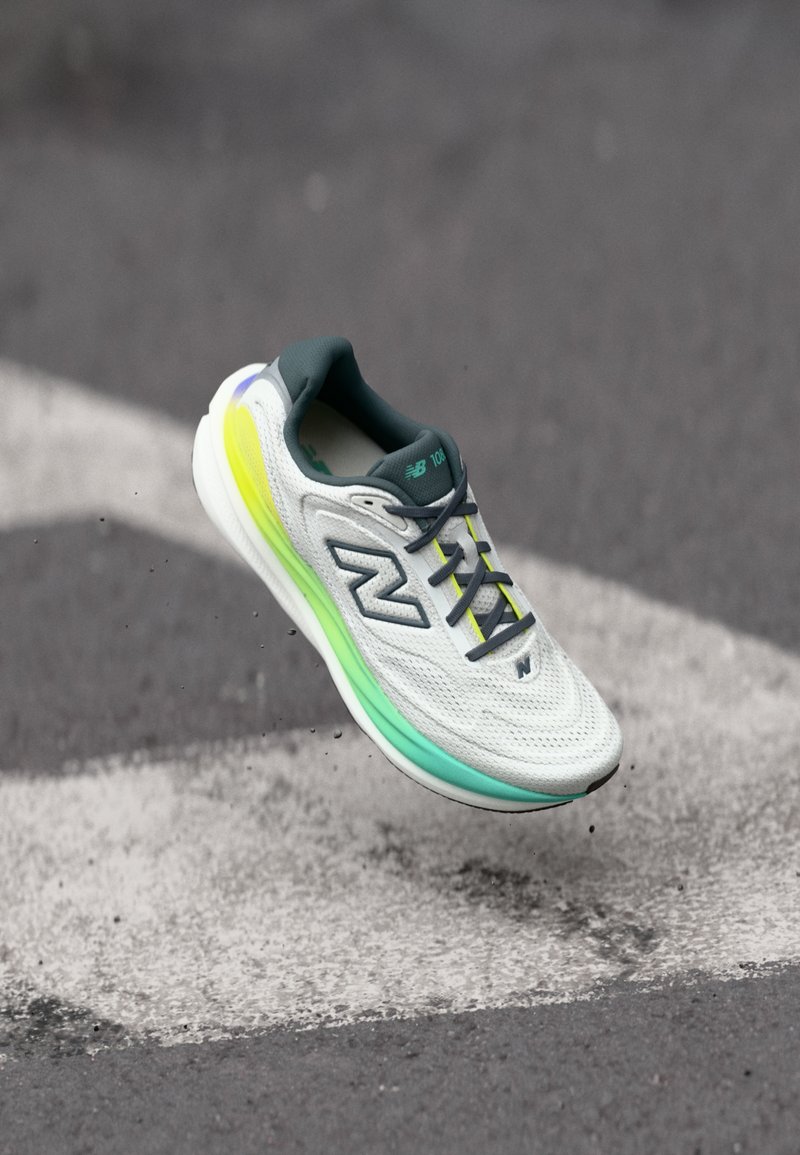 Weißer und türkisfarbener Sportschuh mit grauen Schnürsenkeln und neongradienter Sohle, der über einer Asphaltstraße mit weißen Markierungen schwebt.