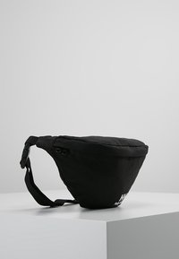 Sac triangulaire noir en tissu durable. Comprend une large ouverture zippée sur le dessus, une sangle réglable et un logo blanc sur le devant.