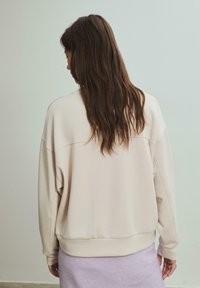 Helle beigefarbene, übergroße Sweatshirt mit rundem Ausschnitt und überschnittenen Schultern. Glatter Stoff, gerippte Bündchen und Saum, getragen über einem lilafarbenen Rock.