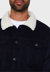 Giacca di velluto a coste blu navy con colletto in finta pelliccia di shearling crema, con due tasche sul petto, chiusura a bottoni e texture a righe verticali.