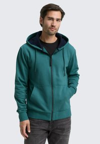 Mann in einer türkisfarbenen Zip-Hoodie mit schwarzem Innenfutter und dunklen Jeans, steht mit einer Hand in der Tasche vor einem einfarbigen Hintergrund.
