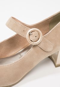 Chaussure Mary Jane en daim beige avec une bride à boucle ronde et un petit talon carré, vue en gros plan sous un angle incliné.