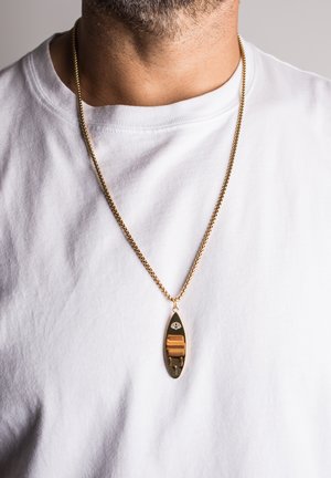 Collier en or avec un pendentif ovale à la surface polie, des détails gravés et une accentuation marron. Porté sur un t-shirt blanc.