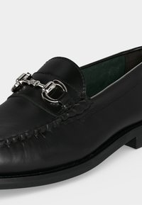 Mocassin en cuir noir avec détail en métal argenté en forme de mors de cheval et dessus cousu, doublure intérieure verte visible et talon bas.