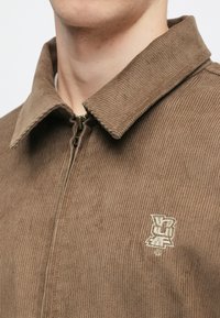 Brun kordfløyelsjakke med glidelås, vid krage og brodert logo på brystet. Teksturert, ribbet stoff med subtil glans.