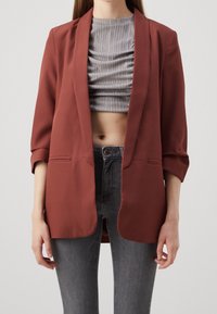 Rust-colored blazer with a smooth texture, open front, and three-quarter sleeves. Worn over a gray, ruched crop top and gray jeans.