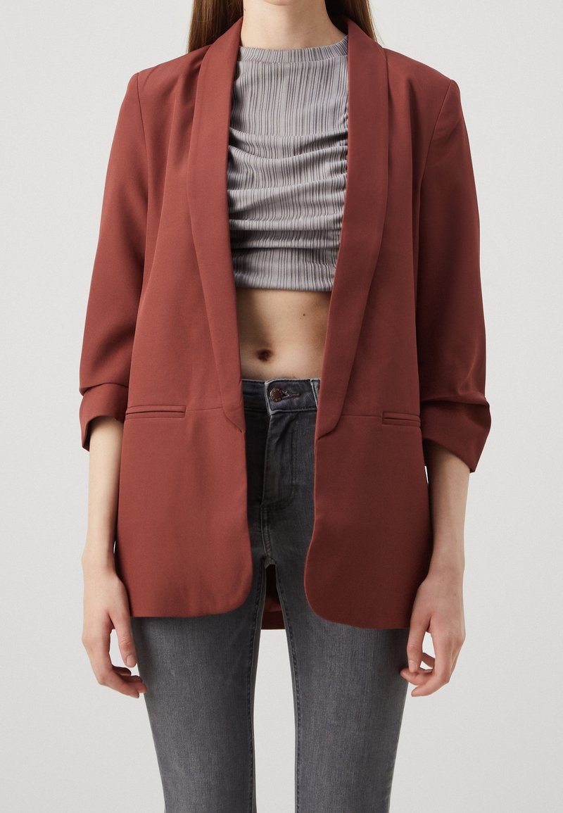 Rust-colored blazer with a smooth texture, open front, and three-quarter sleeves. Worn over a gray, ruched crop top and gray jeans.