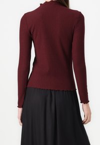Haut long à manches ribbées bordeaux avec un col et des poignets légèrement effilochés, associé à une jupe noire, mettant en valeur un tissu texturé et une coupe ajustée.