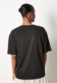Camiseta de algodón negra, de gran tamaño, con cuello redondo, mangas cortas y un acabado desgastado y texturizado. Detalles de costura visibles en los hombros.