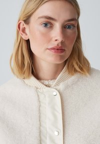 Veste en polaire crème avec un panneau avant lisse beige. Dotée d'un col en côtes, d'une fermeture à boutons-pression et d'une texture duveteuse.