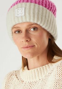 Gebreide beanie met roze en witte strepen, gemaakt van zacht, textuur materiaal, met een decoratief label aan de voorkant.
