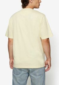 Világos sárga, rövid ujjú póló kerek nyakkivágással. Sima texture, laza fazon. Világos kék farmernadrággal viselve.