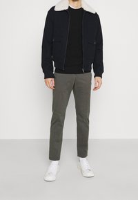 Mężczyzna w czarnej kurtce z białym kołnierzem z owczej wełny, czarnej koszuli, szarych spodniach, białych skarpetach i białych trampkach, stojący na jednolitym tle.