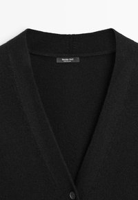 Czarny kardigan wykonany z miękkiej dzianiny, z dekoltem w kształcie litery V i pojedynczym zapięciem na guzik z przodu. Etykieta widoczna wewnątrz.