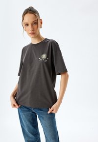 Grijze katoenen t-shirt met een relaxte pasvorm, voorzien van een zonafbeelding en tekst op de linkerborst. Gecombineerd met blauwe spijkerbroek.
