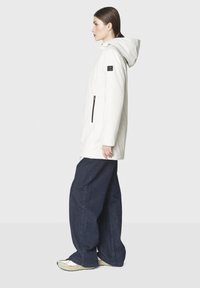 Valkoinen huppujakku, jossa on vetoketjullinen tasku, yhdistettynä rentoihin tummiin farkkuihin ja vaaleisiin kengät, esiteltynä yksinkertaista taustaa vasten.