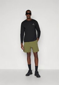 Chemise noire à manches longues avec logo, short vert olive, chaussettes noires et chaussures de sport noires. Le mannequin porte des lunettes de soleil, debout en position neutre sur un fond blanc.