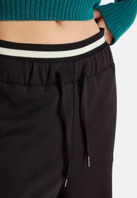 Czarne dresy z elastycznym paskiem w talii, z białym paskiem, sznurek ze metalowymi końcówkami oraz gładką, miękką fakturą.