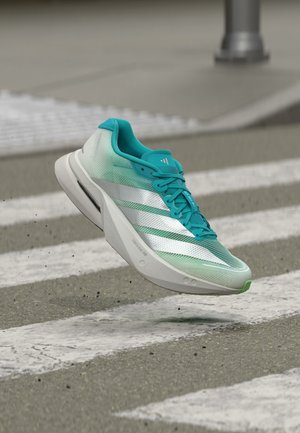 Lichtgewicht groene en witte hardloopschoen met witte strepen en veters, zwevend boven een zebrapad in een stadse straat.