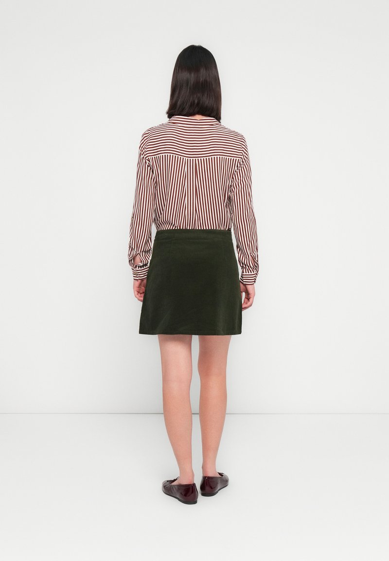 Chemise à manches longues rayée rouge et blanche, associée à une mini-jupe en velours côtelé verte. Le mannequin porte des ballerines bordeaux, sur un fond blanc uni.