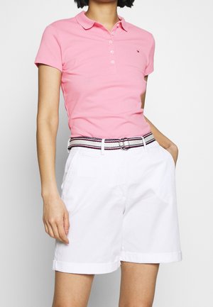 Vrouw draagt een roze polo met korte mouwen, ingestopt in witte short met riem, met een hand in de zak, tegen een effen achtergrond.