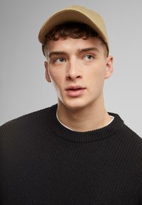 Light brown cap with a curved brim over a black, textured knit sweater. Neutral background highlights the subject.