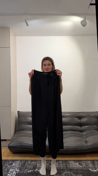 Kvinde står barfodet indendørs og holder et par sorte bukser med høj talje op foran en grå lædersofa mod en hvid væg.