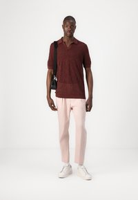 Polo bordeaux à manches courtes avec un motif texturé, associé à un pantalon tapered rose clair. Le modèle porte des baskets blanches et tient un petit sac noir.