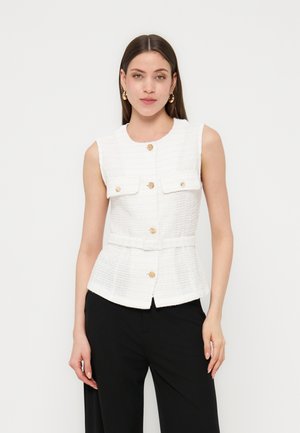 Mujer con chaleco blanco texturizado sin mangas con botones dorados y pantalones negros, posando contra un fondo claro y liso.