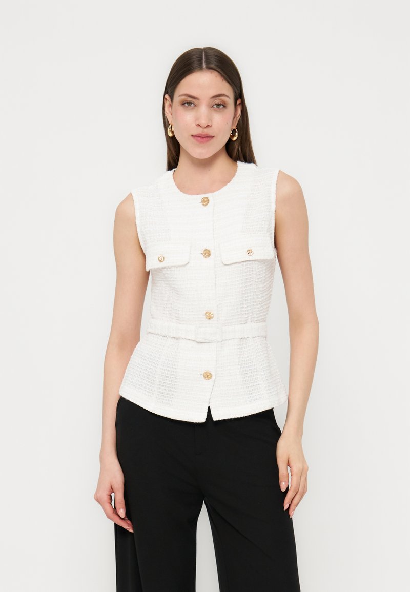 Γυναίκα που φοράει λευκό αμάνικο γιλέκο με υφή και χρυσά κουμπιά, καθώς και μαύρο παντελόνι, στέκεται μπροστά σε ένα απλό φωτεινό φόντο.